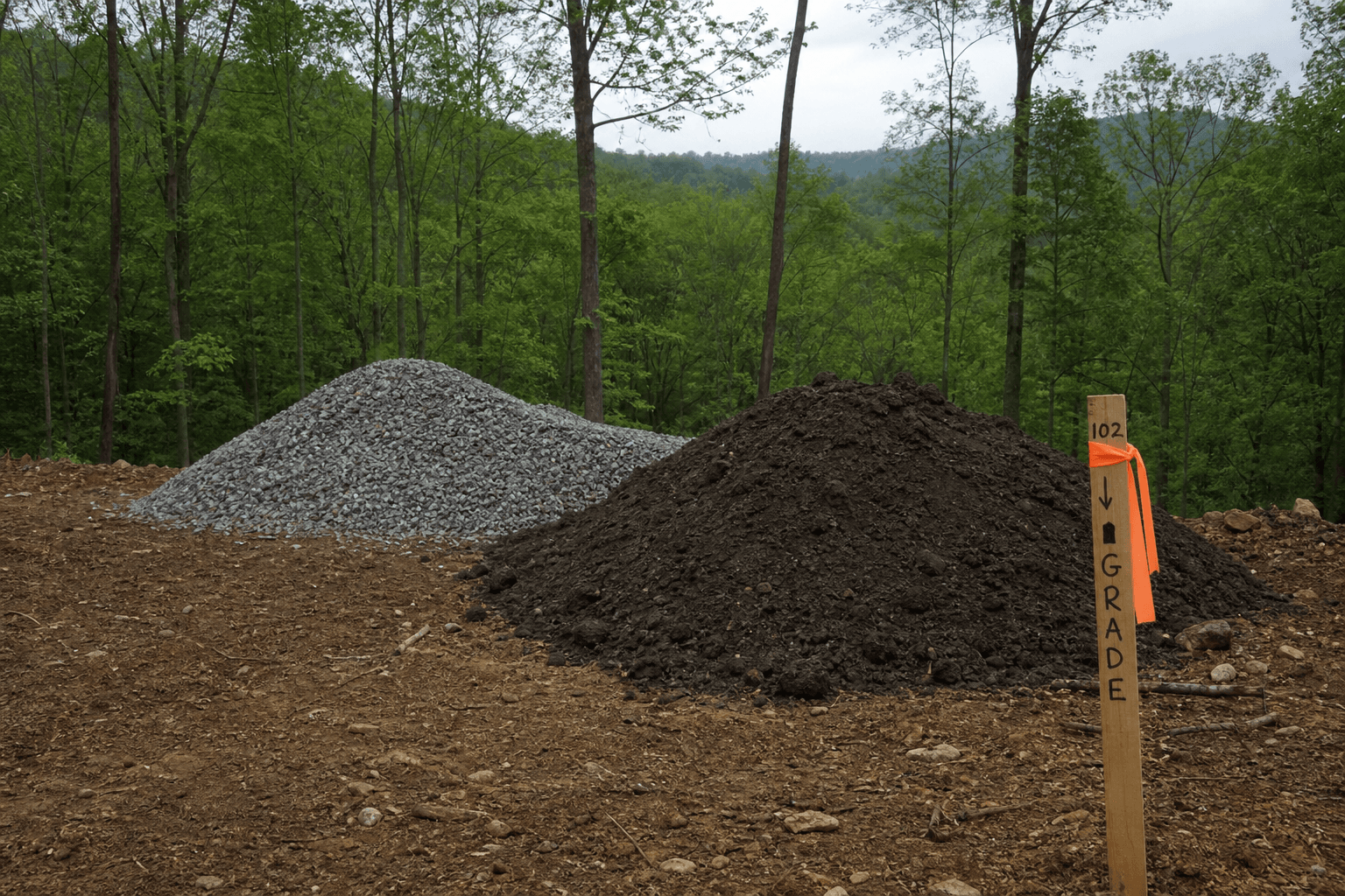 Topsoil and gravel stockpiles next to a grade stake on a sloped restoration lot